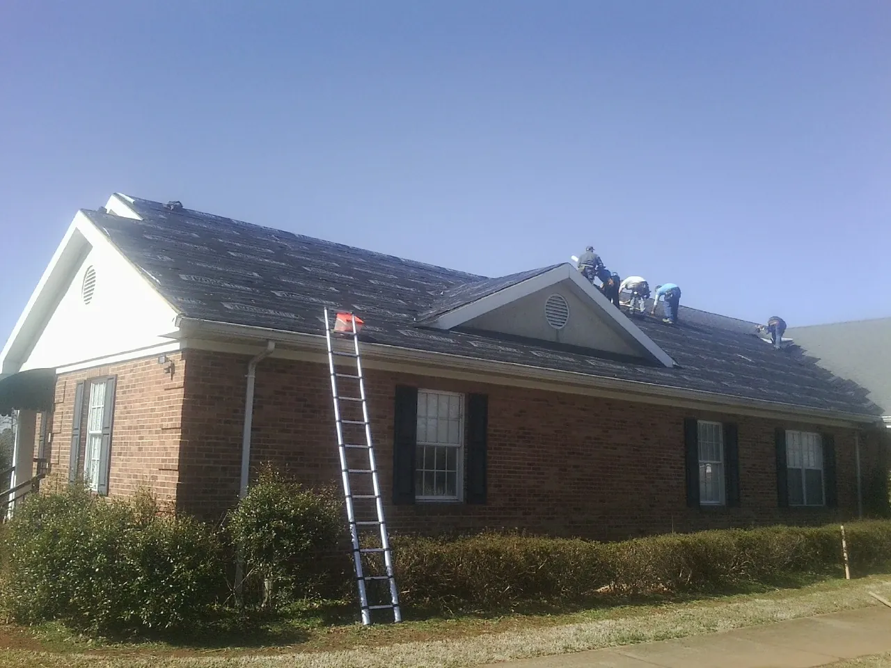 Crew tearing shingles off a brick ranch roof with a ladder set — Metal Roof Repair work by our crew in Blair
