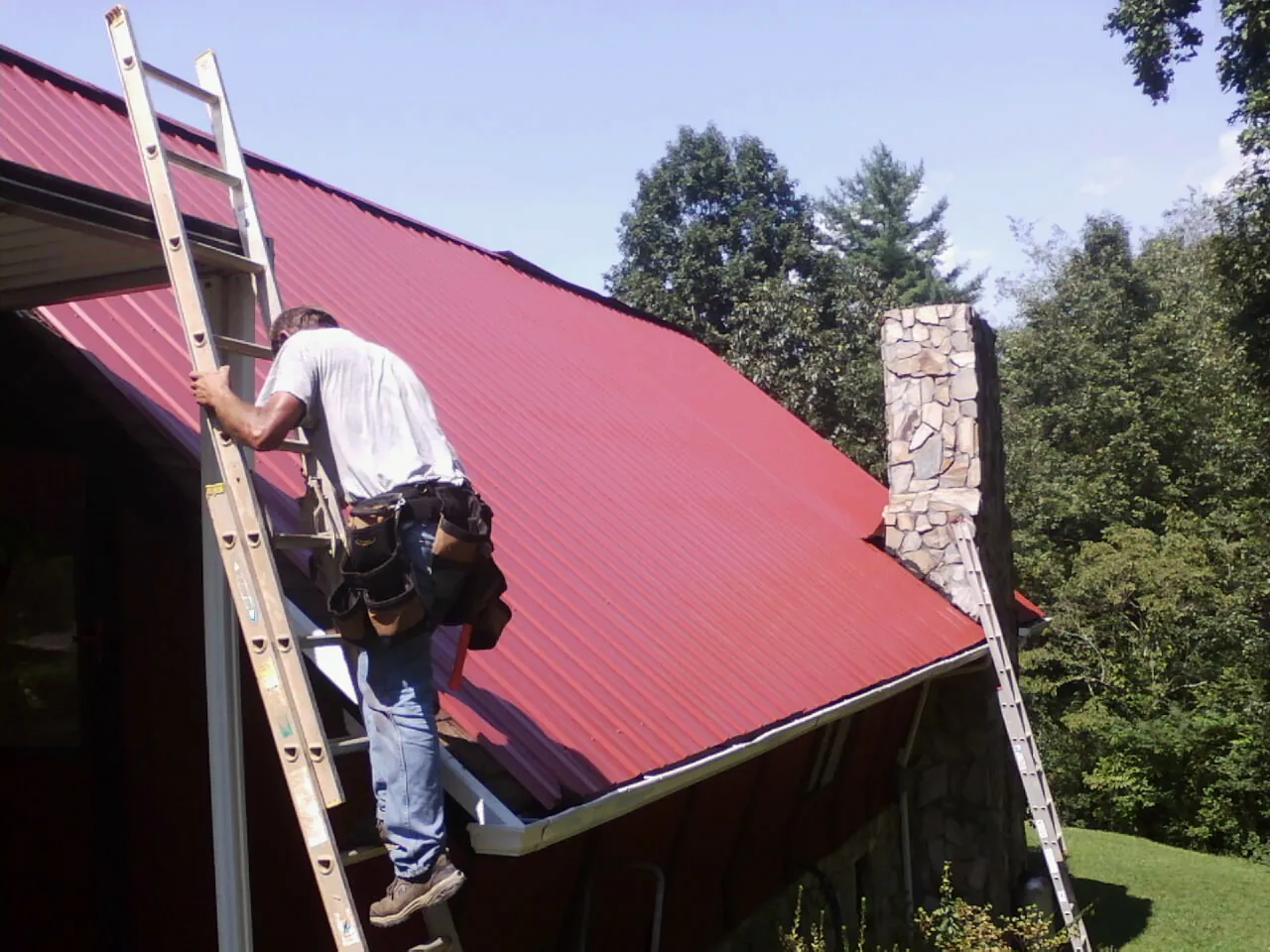 Roofer on a ladder beside a red metal roof and stone chimney — Soffit & Fascia Repair by Roofing Brothers in Blair